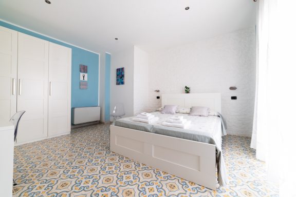 Stanza luminosa con letto matrimoniale e pavimento decorato. Mobili chiari e pareti blu.